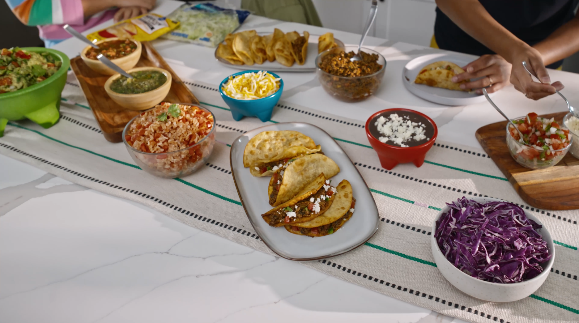 Table with various Mexican dishes including tacos, salads, and sides on a white tablecloth.